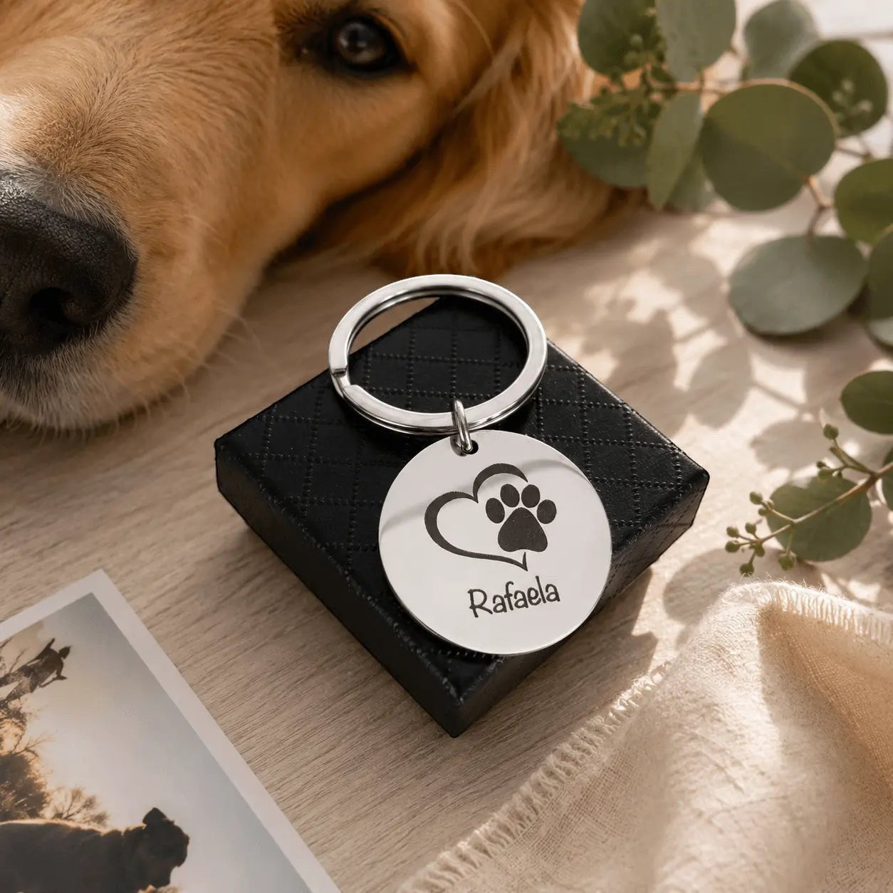 Llavero Moment de acero plateado con grabado de huella y nombre, junto a un perro golden retriever en una mesa