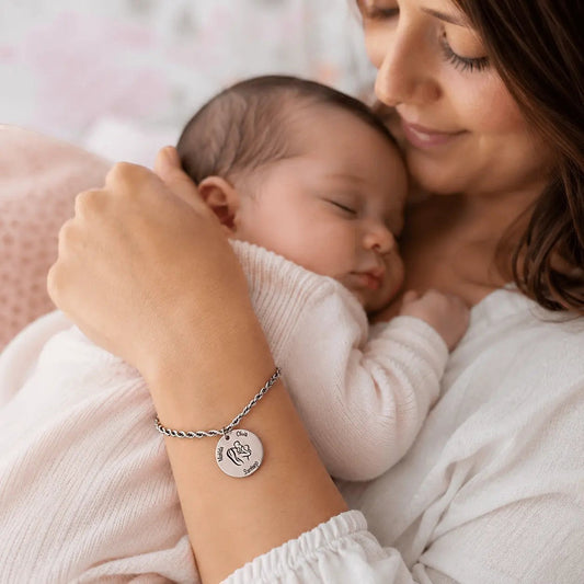 Pulsera Moment de acero inoxidable con medalla grabada personalizada, lucida por una madre con su bebé en brazos.