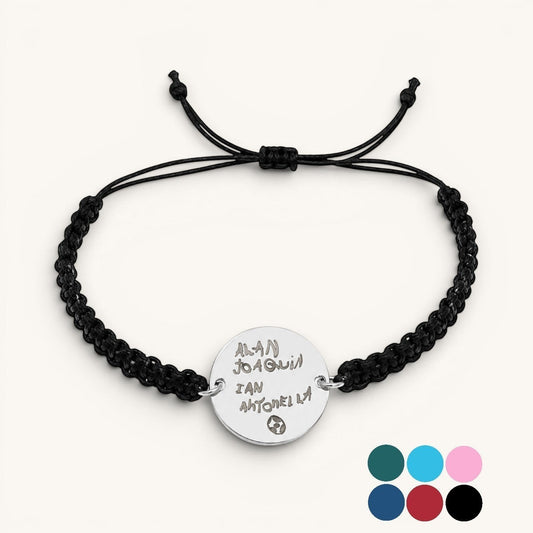 Pulsera Moment de cordón negro ajustable con medalla redonda de plata grabada con nombres y un pequeño dibujo.